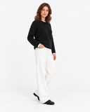 Black round-neck knit sweater shown in a three-quarter pose with white trousers.